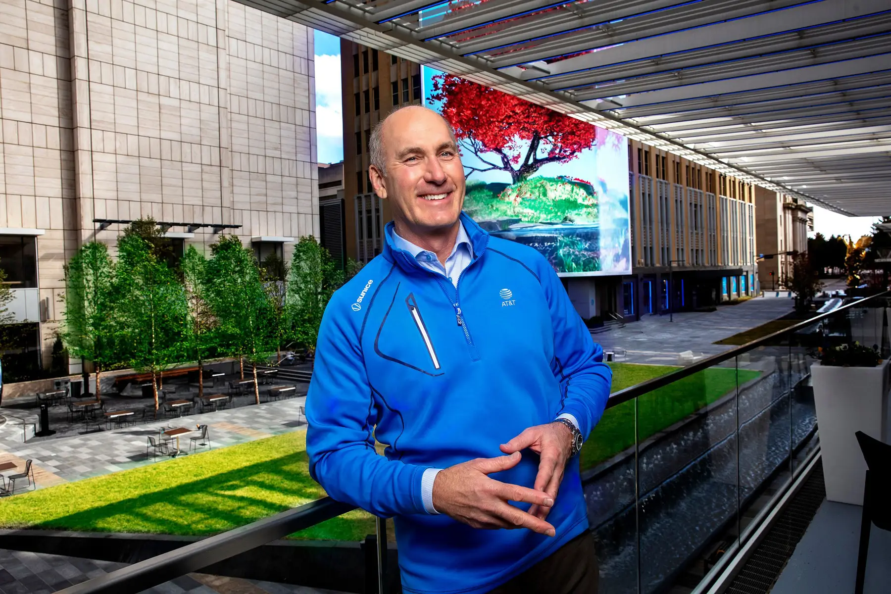 Smiling Man in a Blue Jacket Stands on a Glass Railed Balcony at a Modern Campus Courtyard with Trees and a Large Digital Billboard Behind Him