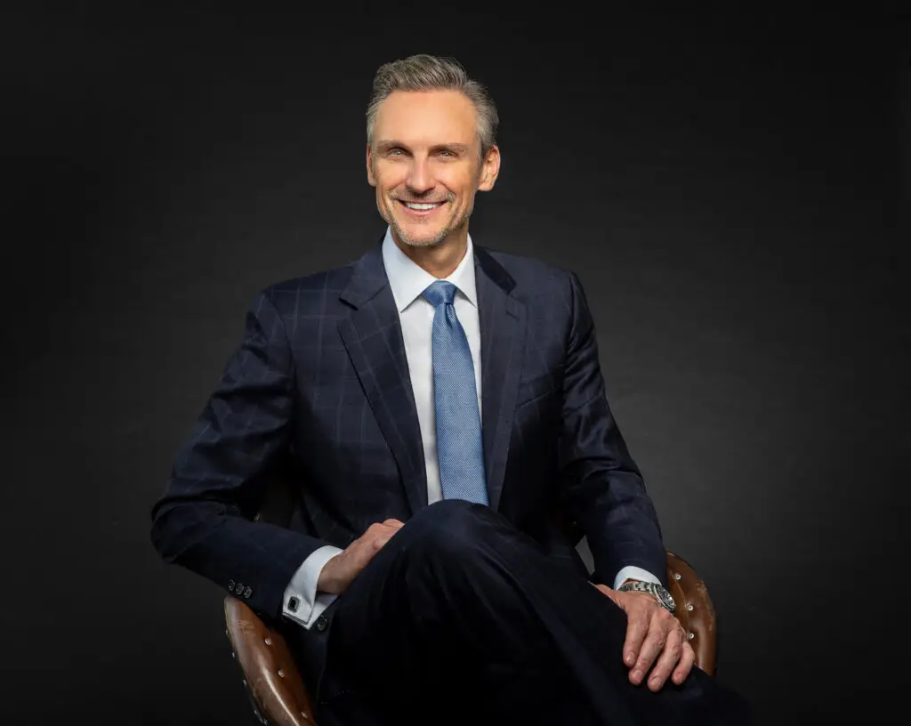 Smiling Businessman in a Navy Check Suit Seated in a Brown Leather Chair Against a Dark Backdrop