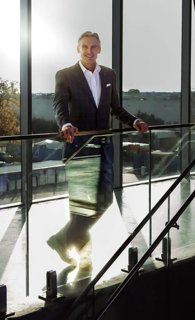 Smiling Businessman in a Dark Checkered Suit Leans on a Glass Railing in a Bright Modern Atrium with Large Windows Behind