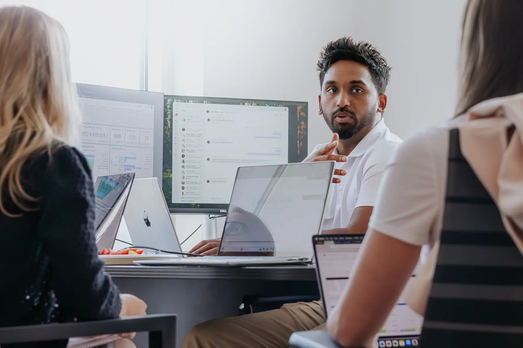 Team Meeting in a Bright Office with Multiple Laptops and Large Monitor Screens Displaying Graphs and a Chat Window While a Man Speaks to Colleagues