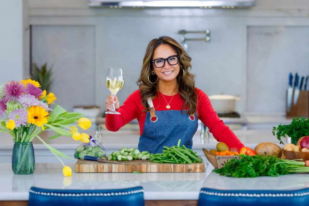 Woman in a Red Shirt and Denim Apron Raising a Wine Glass in a Bright Kitchen with Fresh Flowers and Vegetables on the Counter Behind Her