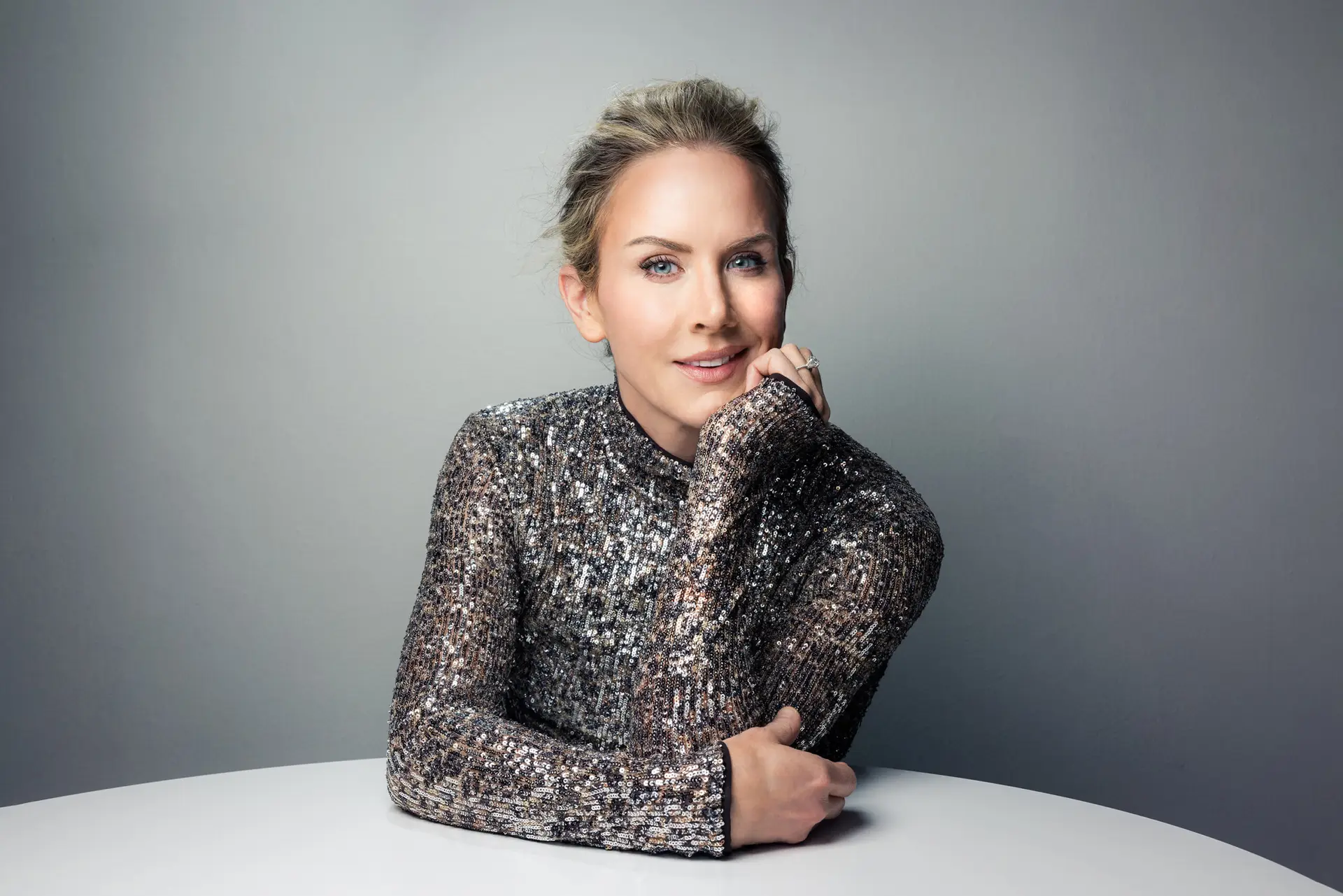 Portrait of a smiling woman in a silver sequin top resting her chin on her hand at a white table.