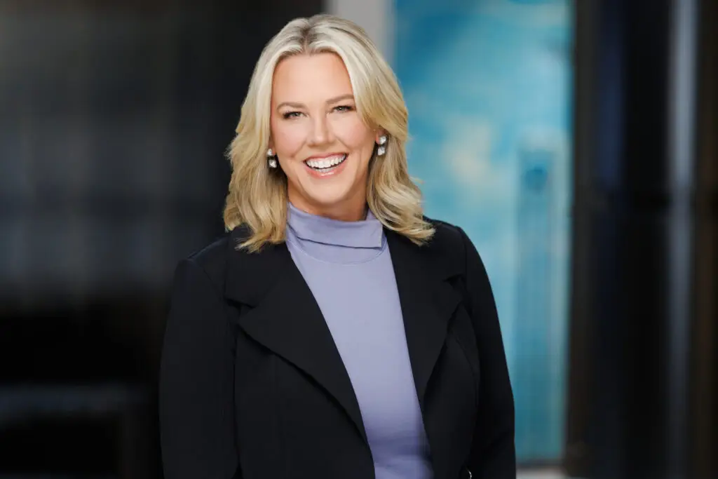 Smiling Blonde Woman in a Black Blazer and Lavender Blouse Head and Shoulders Portrait Against a Blurred Blue Background
