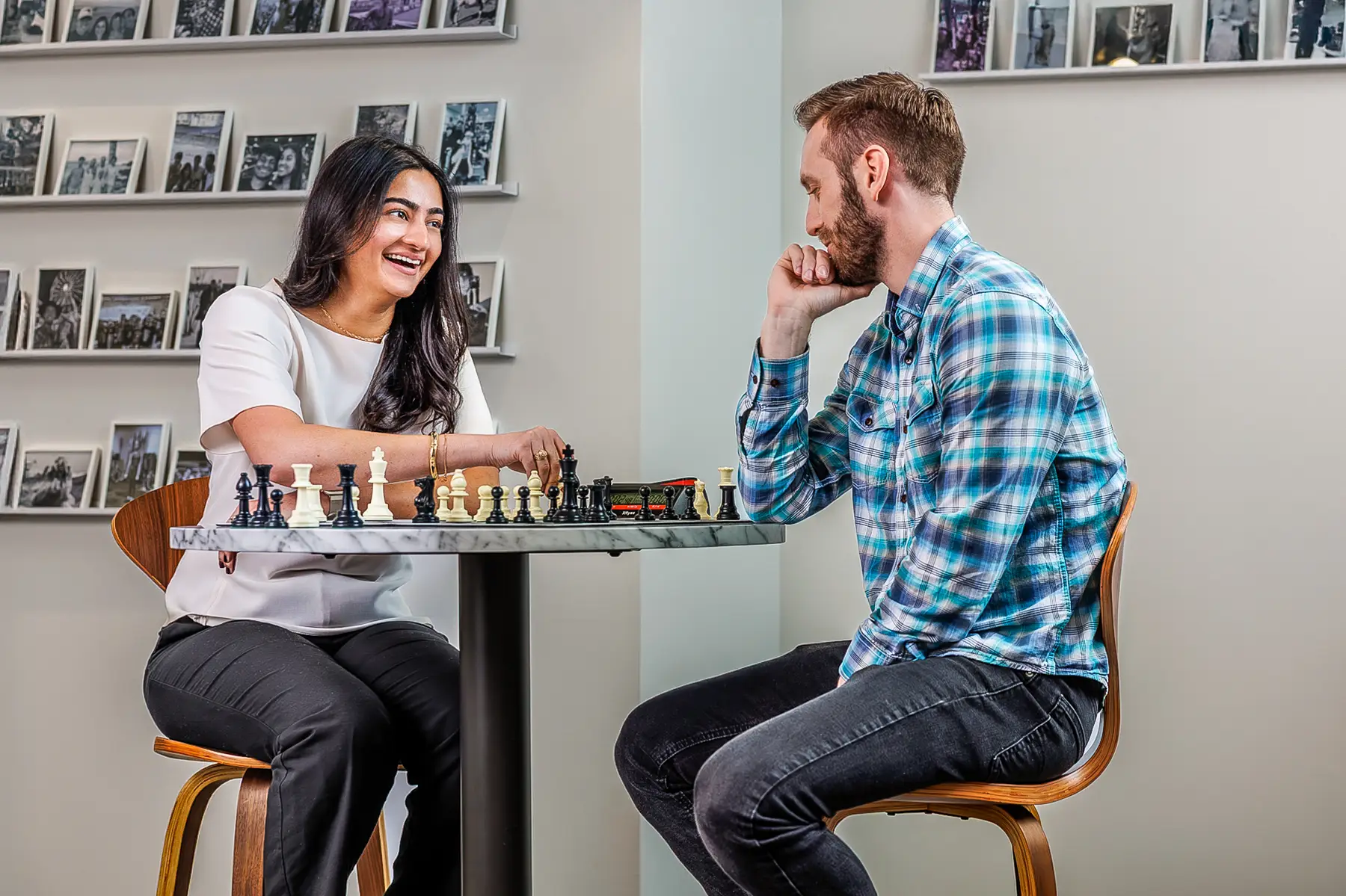 Two Adults Play Chess at a Round Marble Table Smiling and Conversing with Photo Shelves in the Background