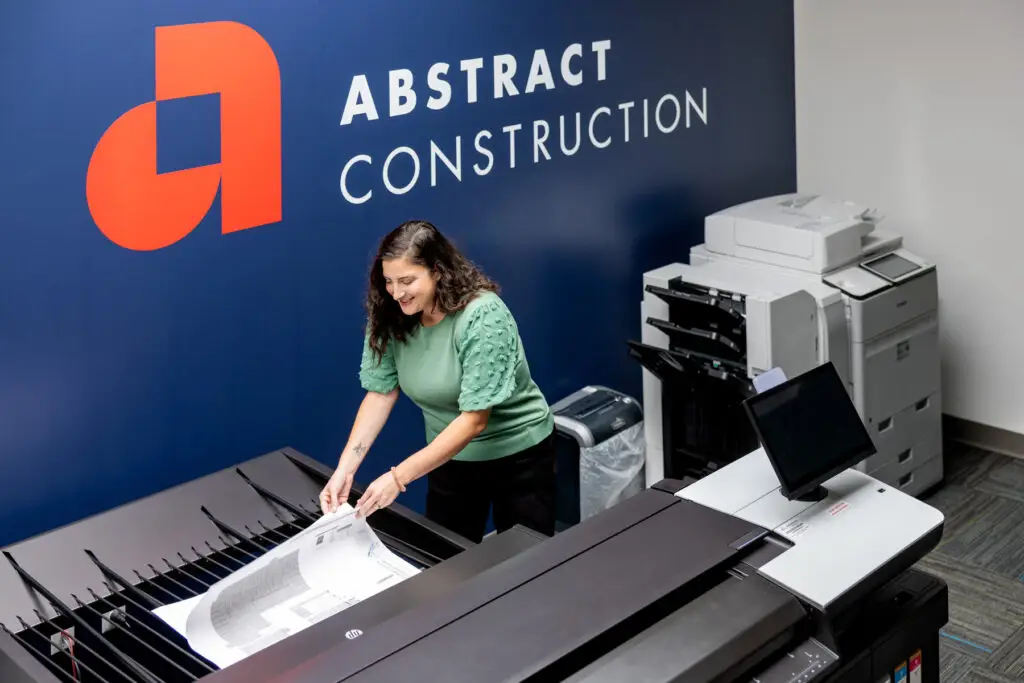 Woman in a Green Top Feeds Papers into a Large Office Copier with a Blue Wall and an Abstract Construction Logo in Orange