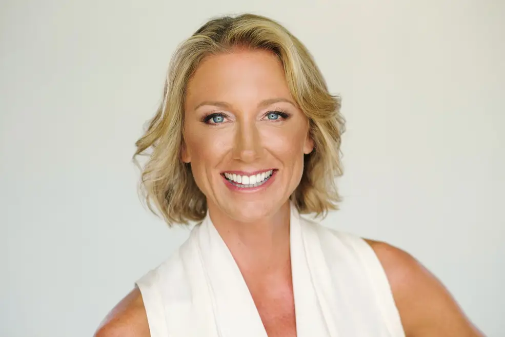 Close Up Portrait of a Smiling Blonde Woman with Blue Eyes in a White Sleeveless Top Against a Light Neutral Backdrop