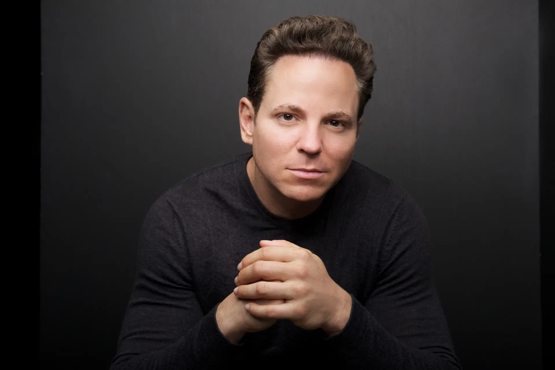 Portrait of a Man with Dark Hair and a Charcoal Sweater Hands Clasped in Front Neutral Expression Against a Dark Backdrop