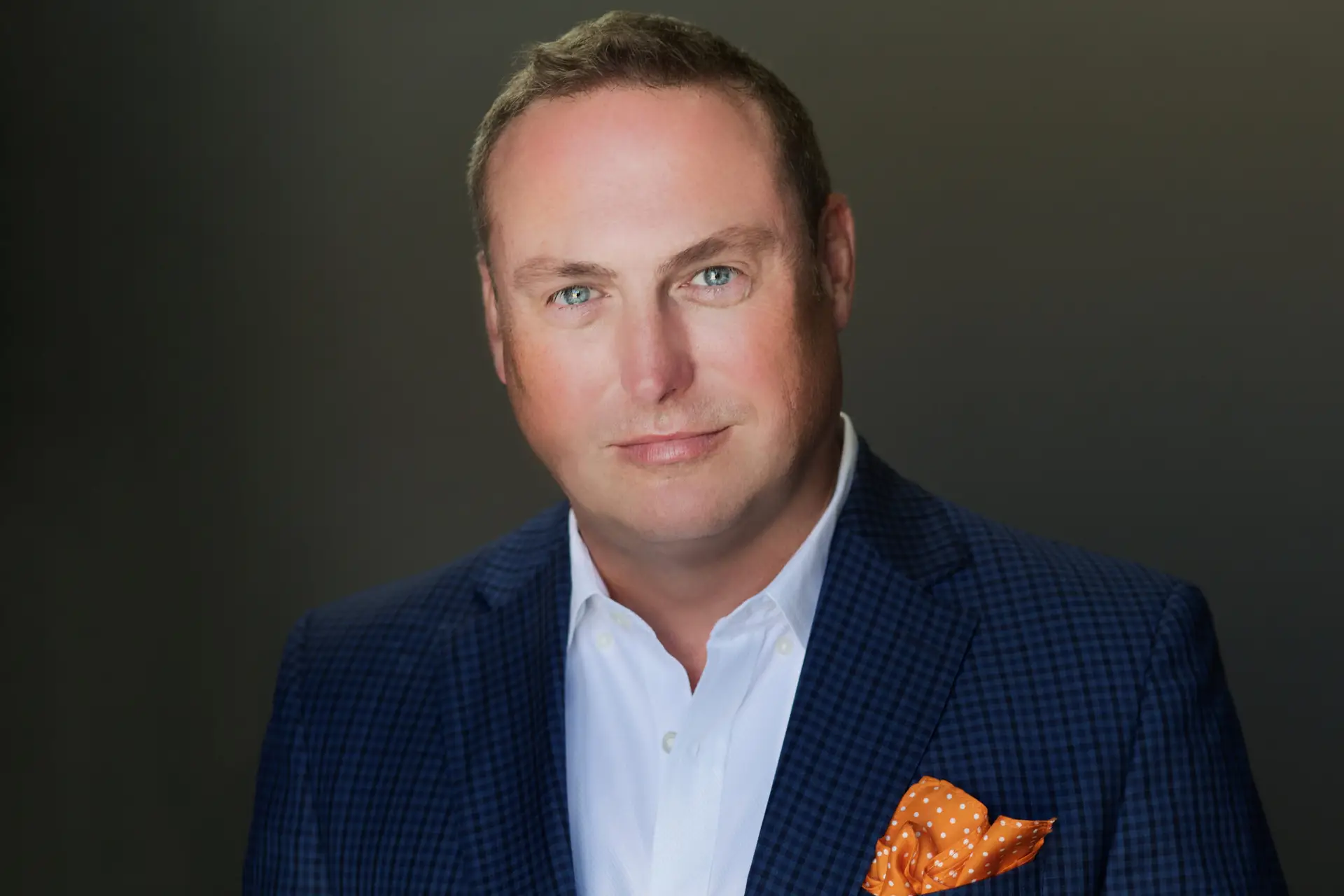 Portrait of a man in a blue check blazer and white shirt with an orange polka-dot pocket square, smiling softly against a dark background and centered in the frame