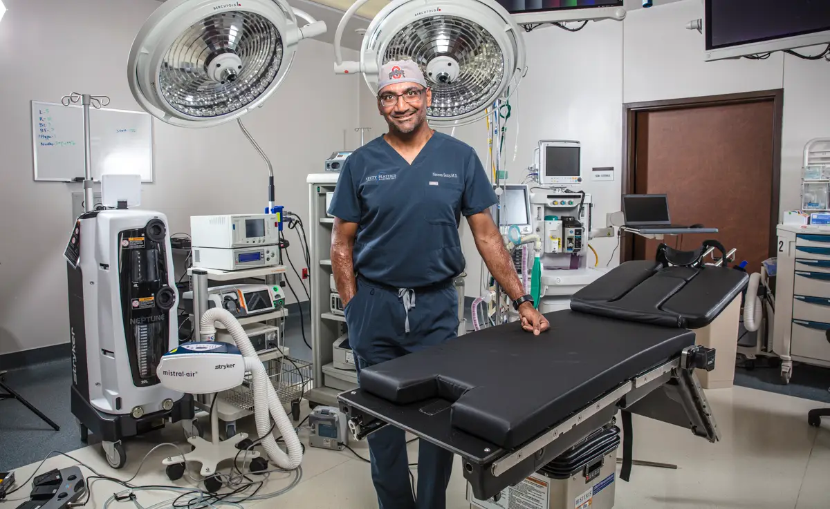 Surgeon Personal Branding Portrait in the Operating Room - Kelly Williams Photography - Kelly Williams Photography Surgeon in Navy Scrubs and Cap Standing Beside an Empty Operating Table in a Bright Well Equipped Hospital Operating Room with Overhead Surgical Lights and Monitors Nearby