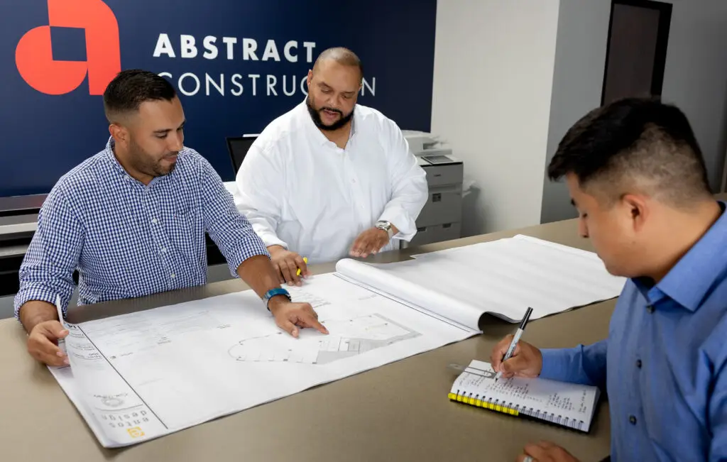 Three Construction Professionals Review Large Architectural Blueprints on a Conference Table in an Office