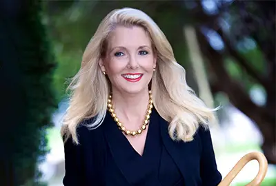 Portrait of a smiling blonde woman in a dark blazer wearing a gold bead necklace, outdoors with greenery in the background.