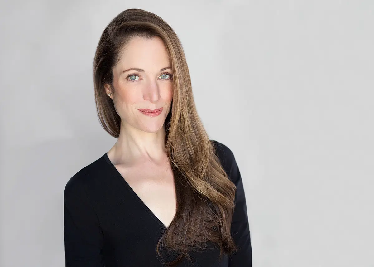 Head and Shoulders Portrait of a Smiling Woman with Long Brown Hair in a Black V Neck Top Against a Neutral Gray Background
