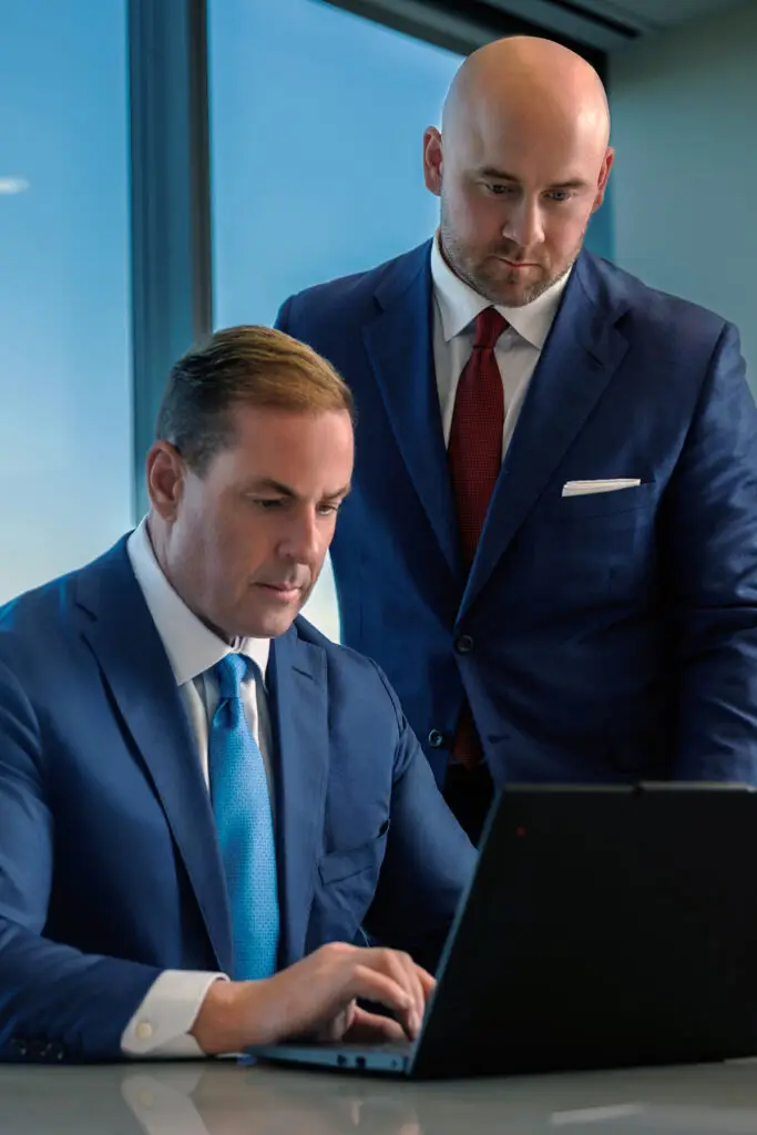 Two Businessmen in Blue Suits Collaborate at a Laptop One Seated Typing While the Other Stands Nearby Reviewing the Screen in a Bright Office